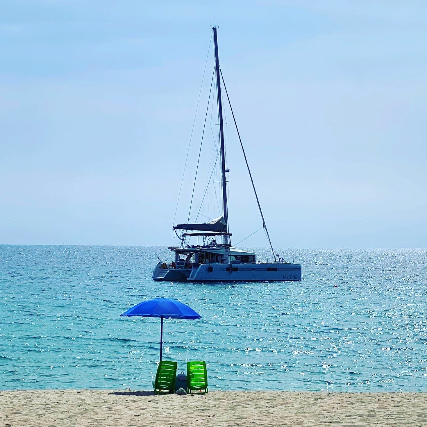 Blauer Sonnenschirm am Strand, dahinter die MY·ONE vor Anker